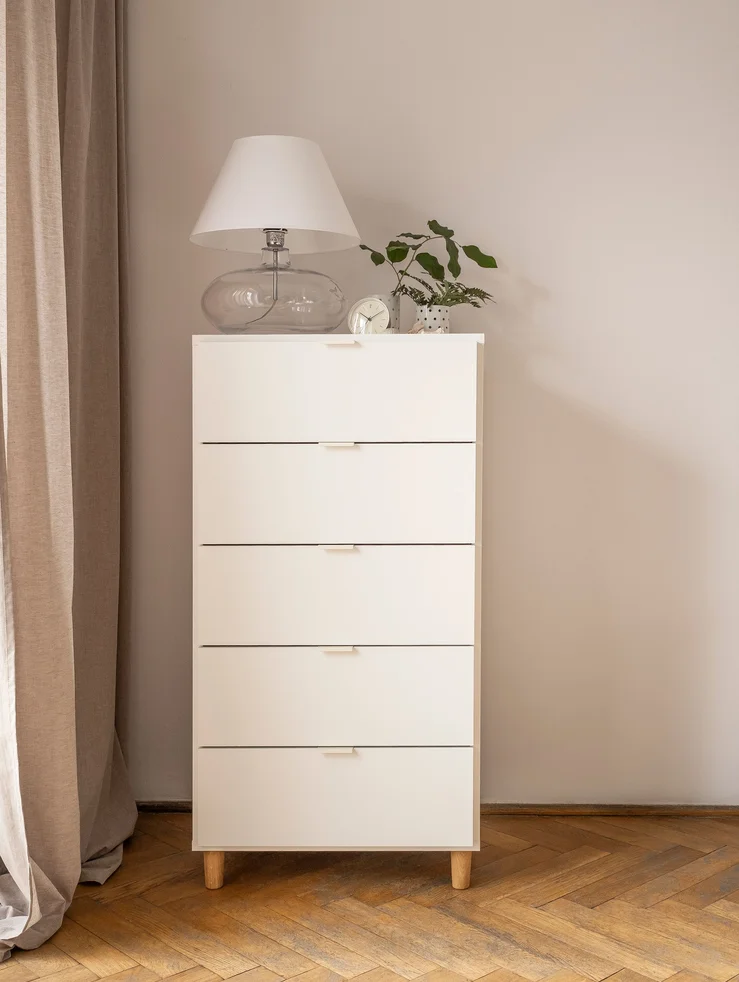 Wysoka biała komoda z lampą i roślinami na jasnej ścianie.